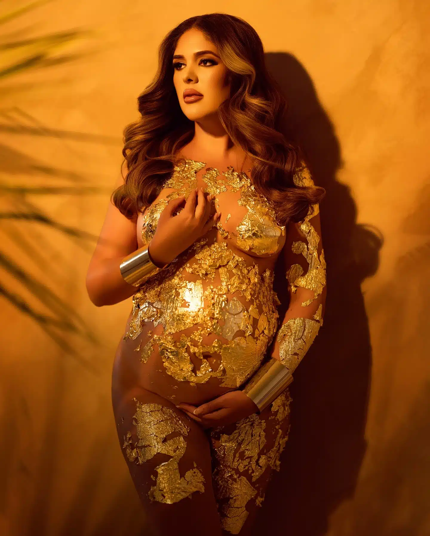 Pregnant woman in gold leaf body art and metallic arm cuffs posing against a warm golden wall with palm shadows at a Mia | Creative Canvas Photo Studio Miami Pregnant woman in gold leaf body art and metallic arm cuffs posing against a warm golden wall with palm shadows at a Miami photo studio