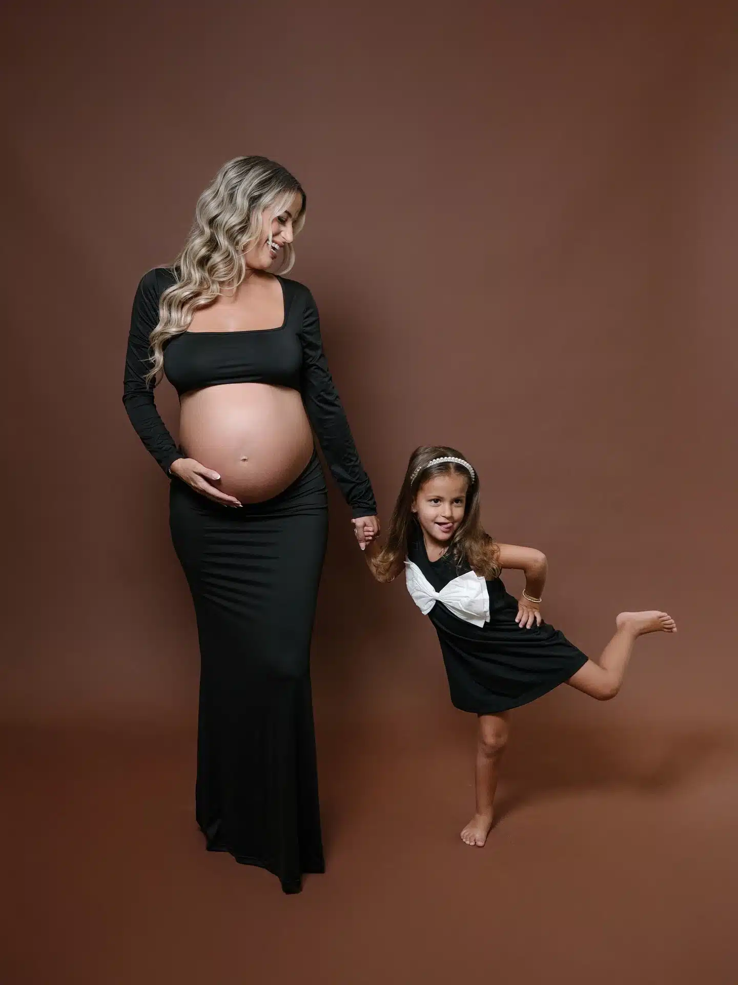 Pregnant mother in a black cutout gown holding hands with her young daughter in a black bow dress on a brown backdrop at | Creative Canvas Photo Studio Miami Pregnant mother in a black cutout gown holding hands with her young daughter in a black bow dress on a brown backdrop at a Miami photo studio