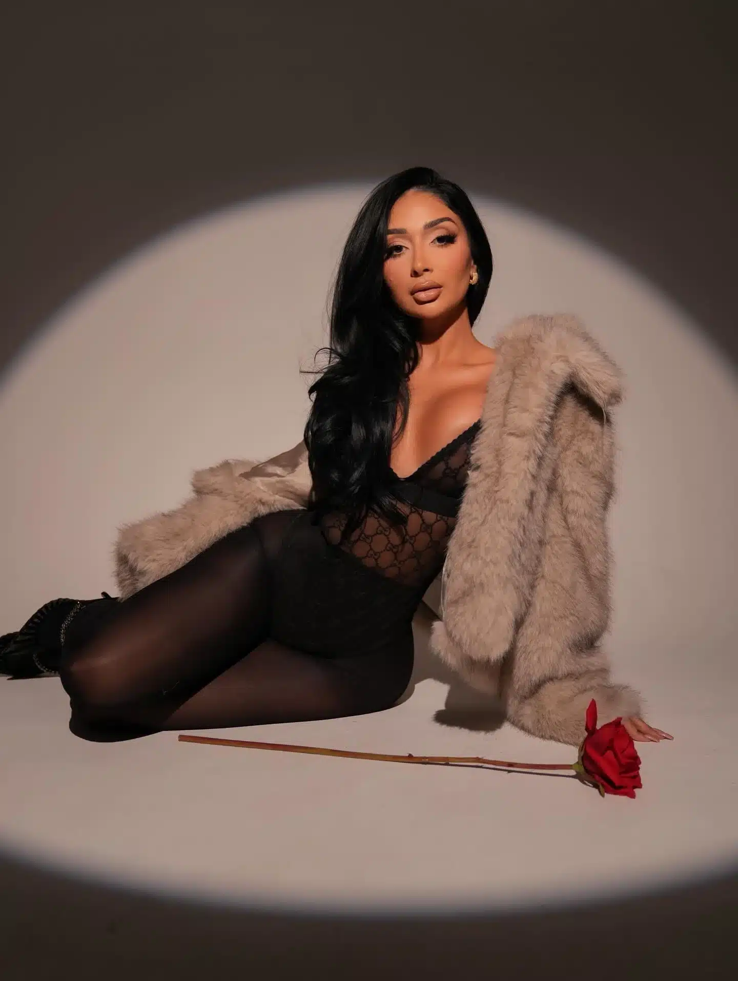 Woman reclining on the floor in a black lace bodysuit and taupe fur coat beside a red rose in a spotlight at a Miami photo studio