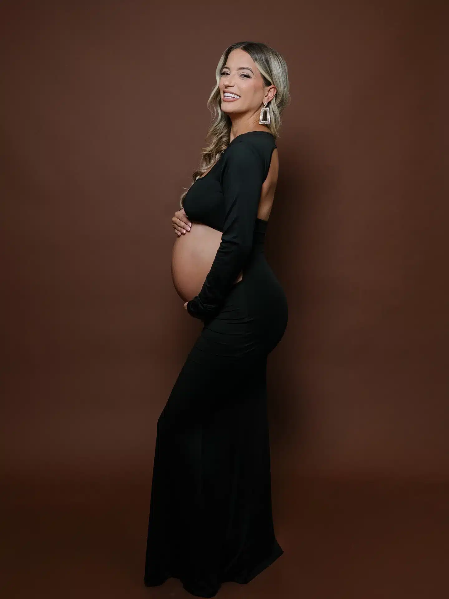 Smiling expectant mother in a black cutout maternity gown with statement earrings holding her bump against a brown backd | Creative Canvas Photo Studio Miami Smiling expectant mother in a black cutout maternity gown with statement earrings holding her bump against a brown backdrop at a Miami photo studio