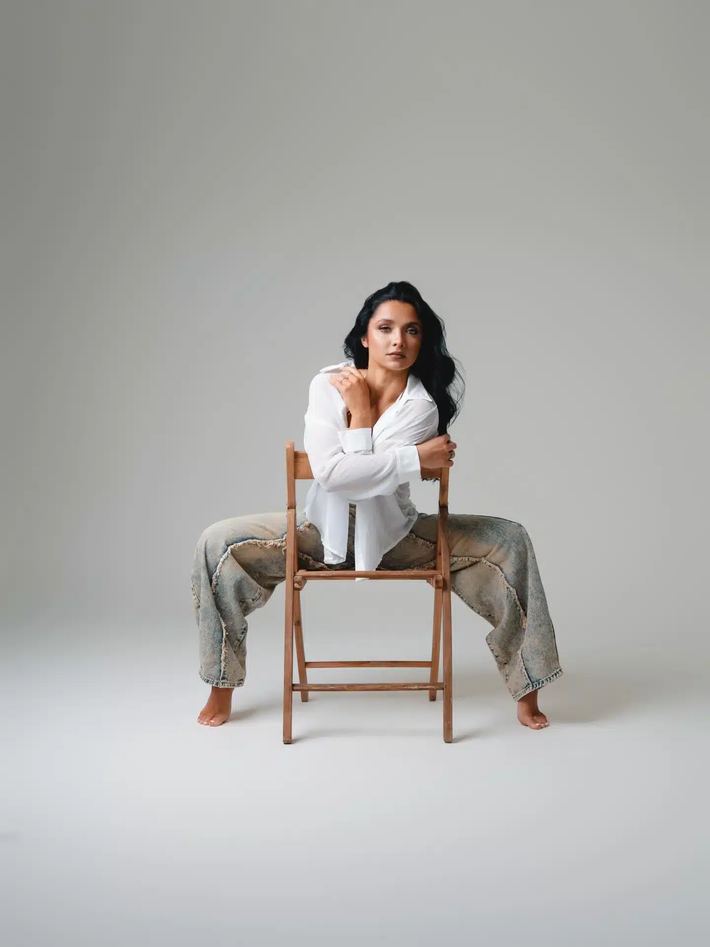 Woman in a white shirt and wideleg distressed jeans seated on a wooden folding chair against a gray backdrop at a Miami | Creative Canvas Photo Studio Miami Woman in a white shirt and wide leg distressed jeans seated on a wooden folding chair against a gray backdrop at a Miami photo studio