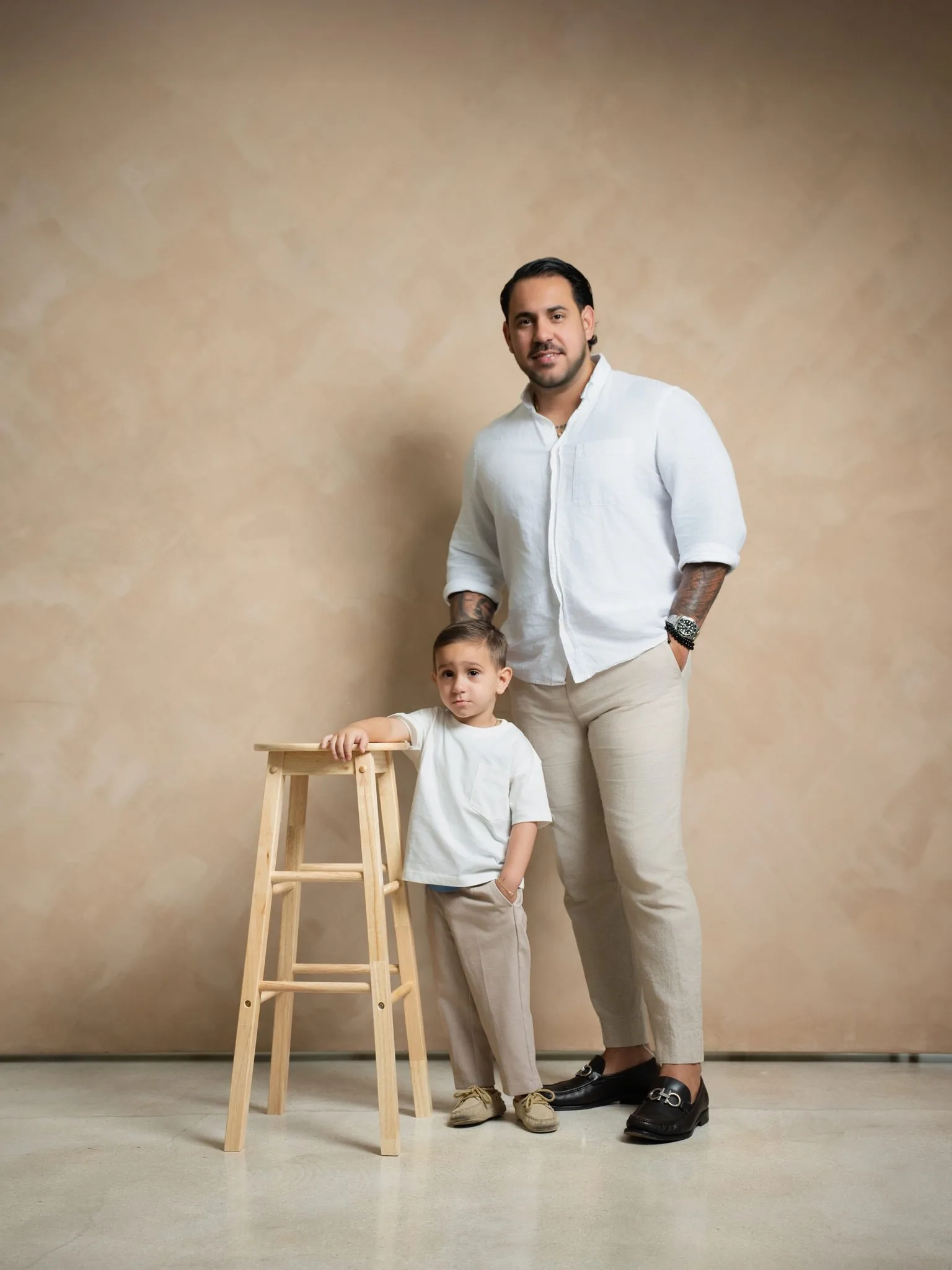 Family Photography by a Miami Photographer | Father and Son | Creative Canvas Photo Studio Miami Sesión de fotos familiar con fotógrafo en Miami papá de blanco y beige con su hijo junto a un taburete sobre un fondo beige