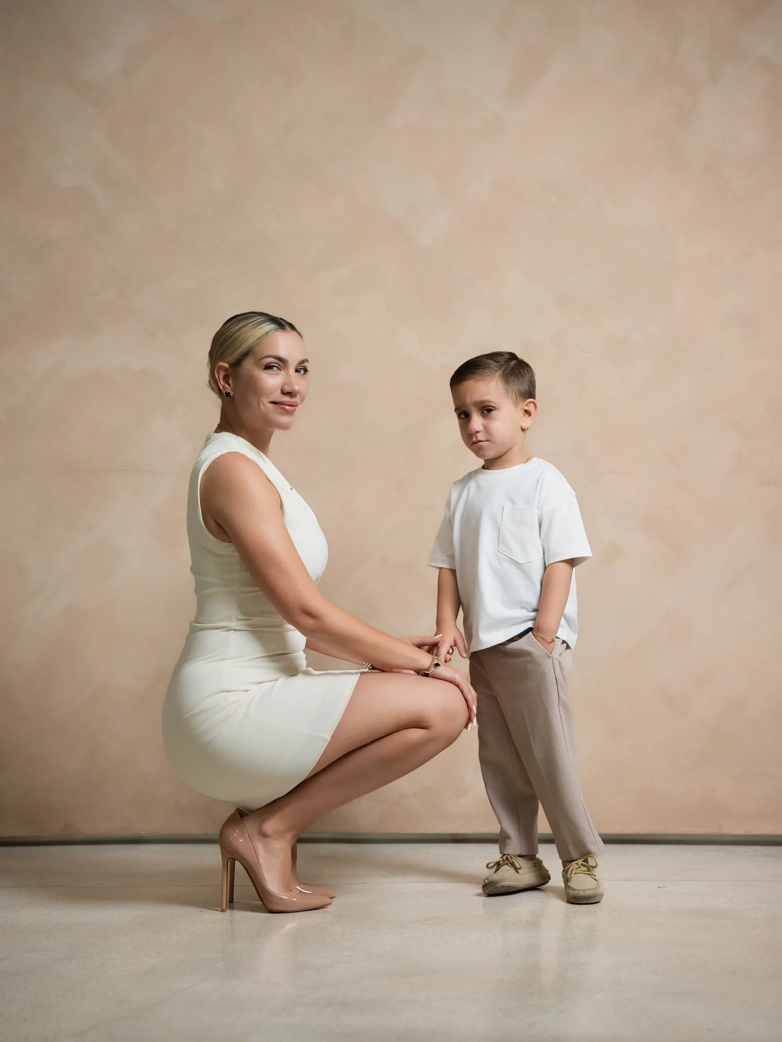 Family Photos Miami Photo Studio | Mother and Son | Creative Canvas Photo Studio Miami Fotos familiares en un estudio de fotos de Miami mamá con vestido crema agachada junto a su hijo sobre un fondo beige