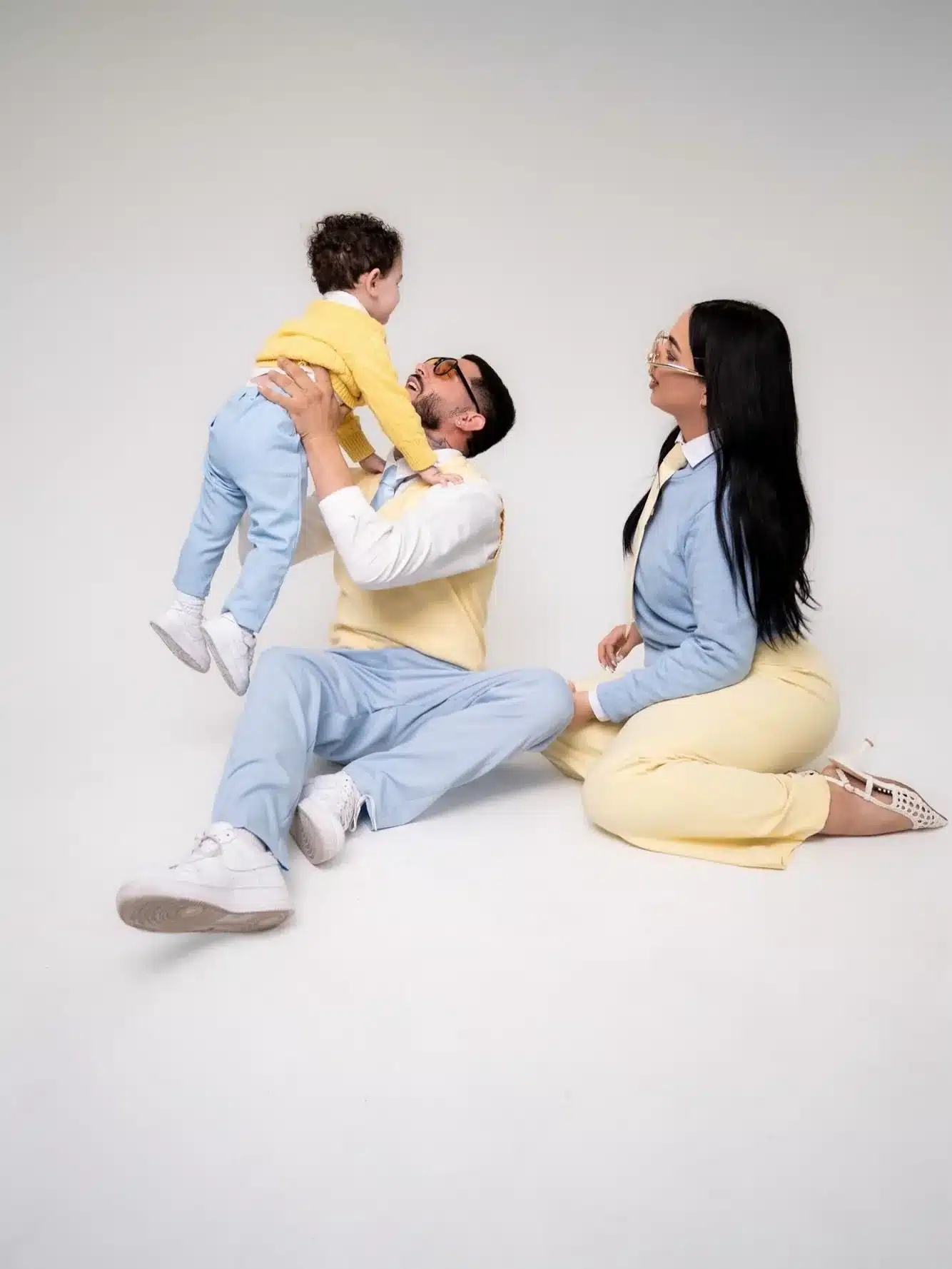 Family Photoshoot Miami Photo Studio | Dad Lifts His Son | Creative Canvas Photo Studio Miami Sesión de fotos familiar en Miami papá levantando a su hijo de suéter amarillo mientras mamá ríe sobre un fondo blanco