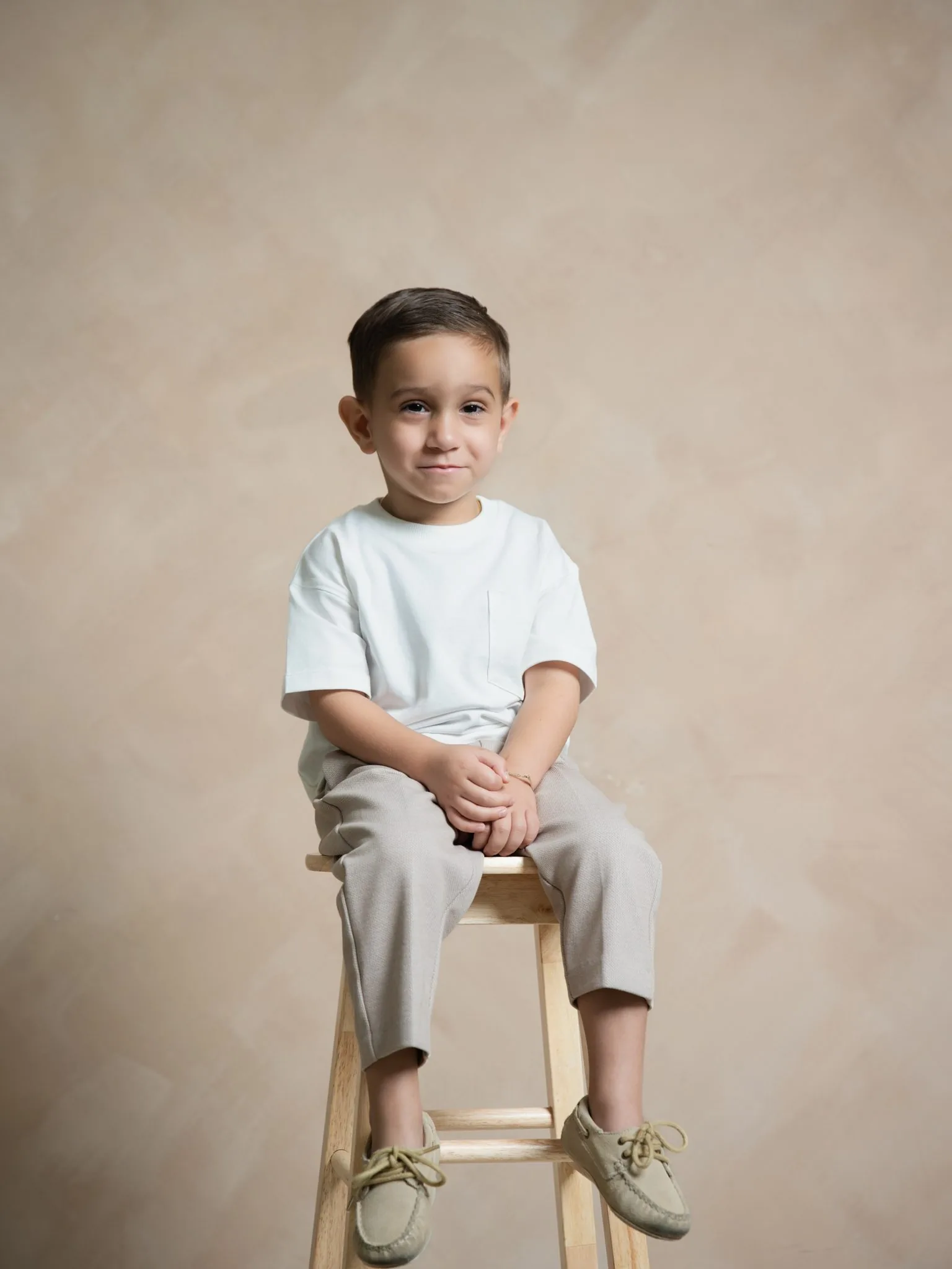 Family Portrait Session Miami Photography Studio | Toddler | Creative Canvas Photo Studio Miami Fotos familiares en un estudio de fotografía de Miami niño con camiseta blanca y pantalón beige sentado en un taburete