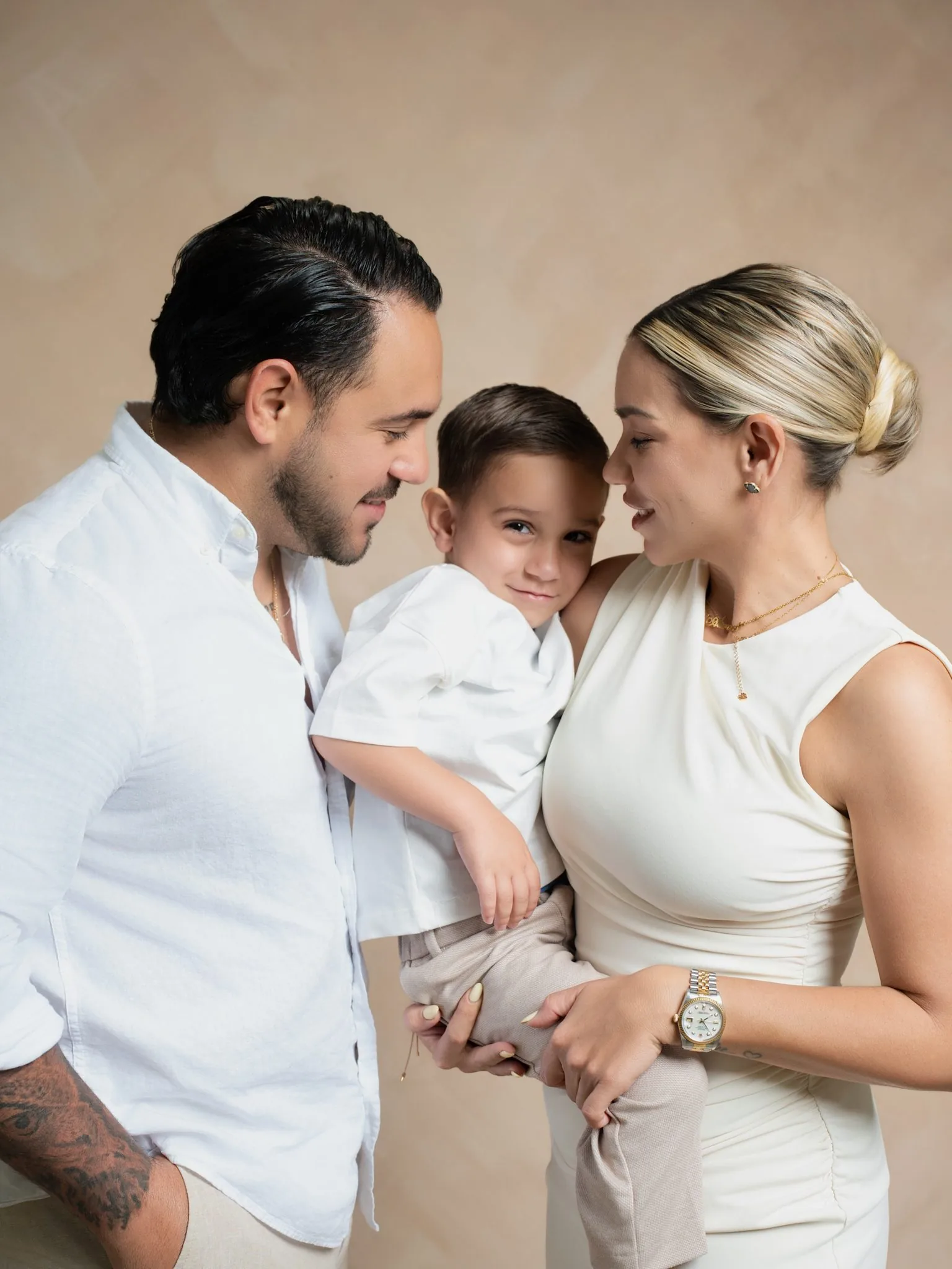 Miami Family Photographer | Parents Hugging Their Son | Creative Canvas Photo Studio Miami Sesión de fotos en familia en Miami padres de blanco y crema abrazando a su hijo sobre un fondo beige